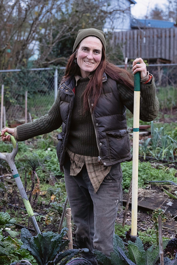 A woman standing out in a garden with gardening tools in hand.